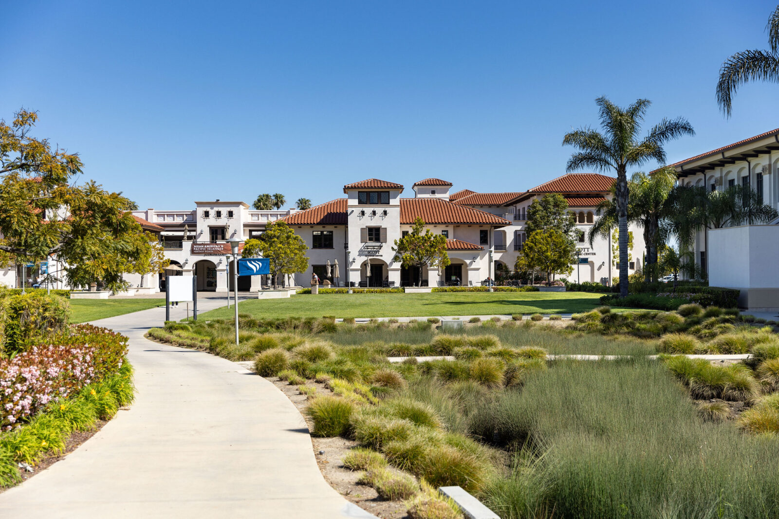 A landscaped outdoor scene with a curved concrete walkway in the foreground, ornamental grasses, palm trees, and a large Spanish-style building with white walls and red-tiled roofs under a clear blue sky.